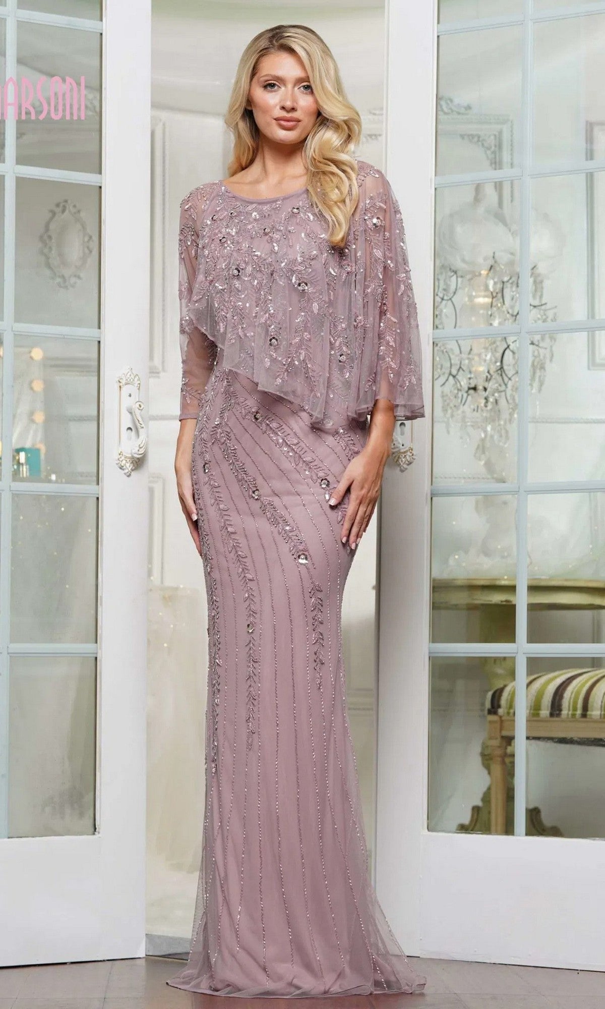 A woman stands indoors in front of glass doors wearing the Marsoni MV1343 long formal dress—a mauve, floor-length column gown with beaded embellishments and a sheer flowing overlay.